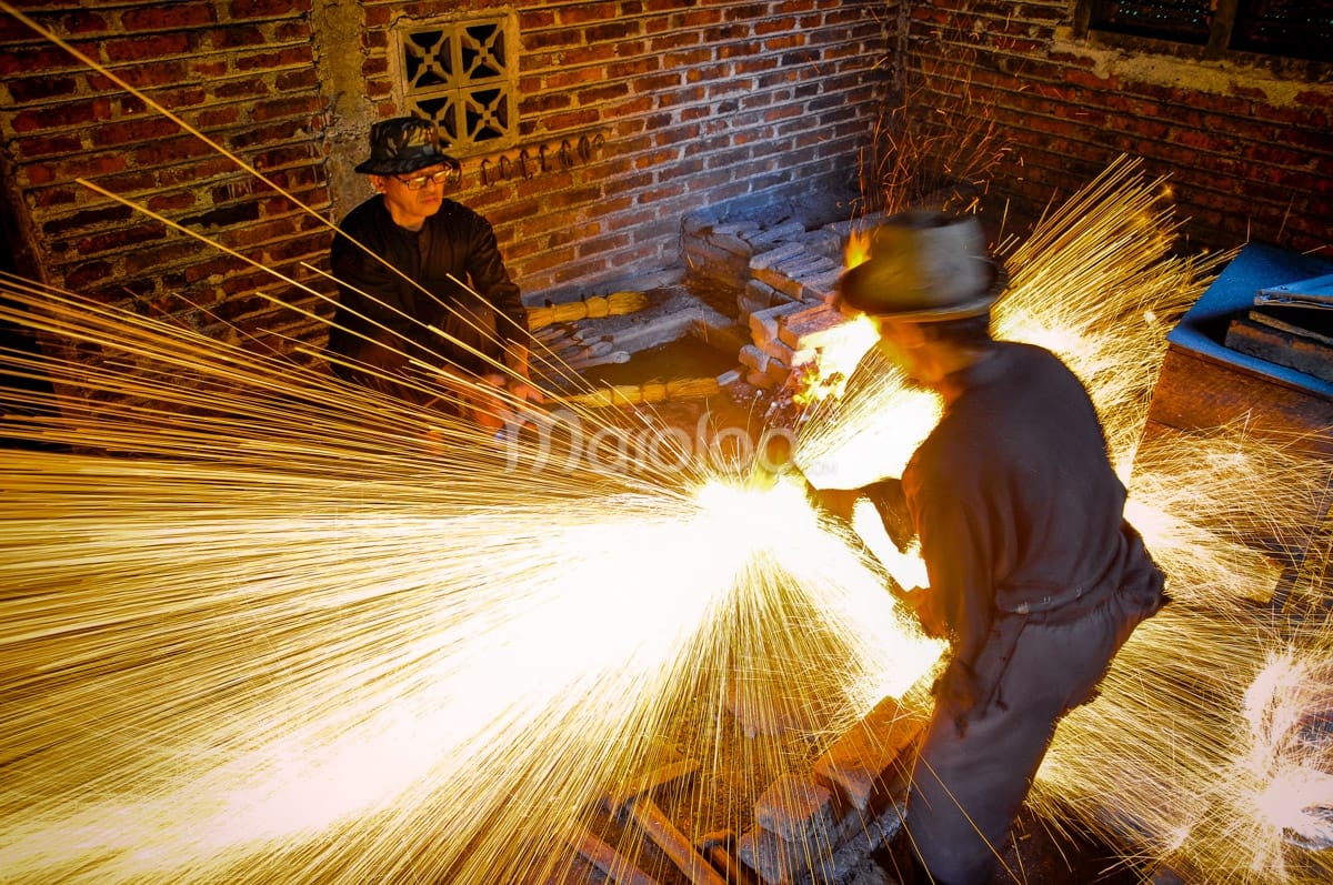 Two craftsmen working in a traditional forge with bright, orange sparks flying as they hammer a keris.