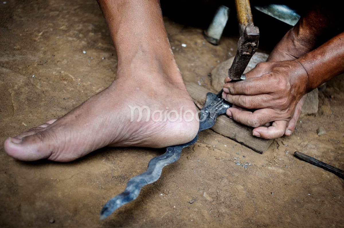 Close-up of a craftsman’s hands and foot shaping the blade of a keris with a hammer.