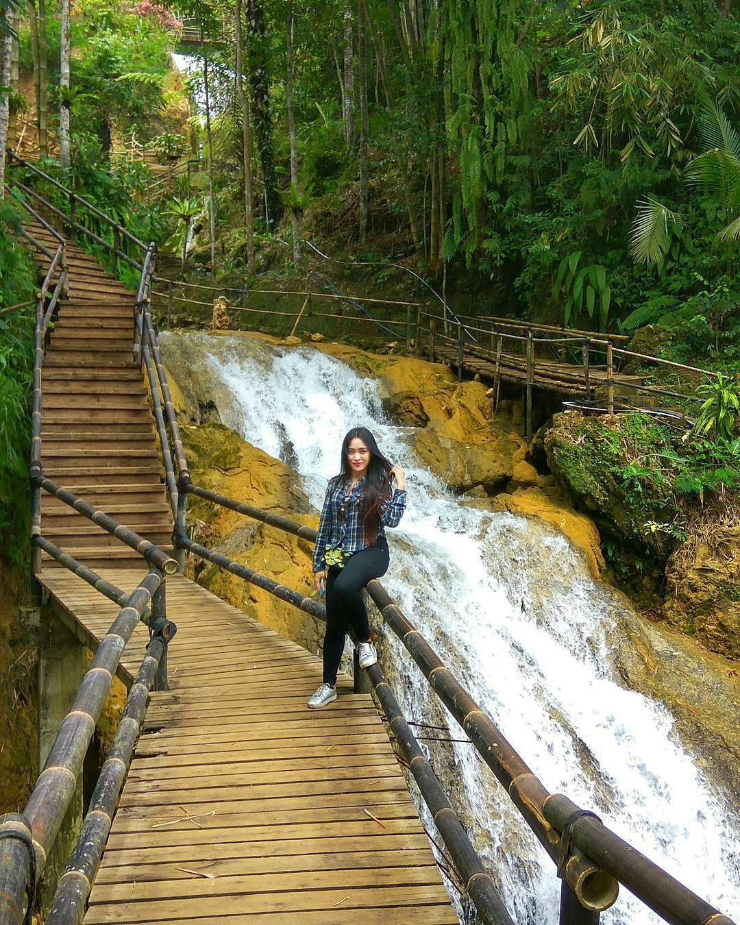 A woman poses on a wooden walkway next to a cascading waterfall surrounded by green trees at Mudal River Park.