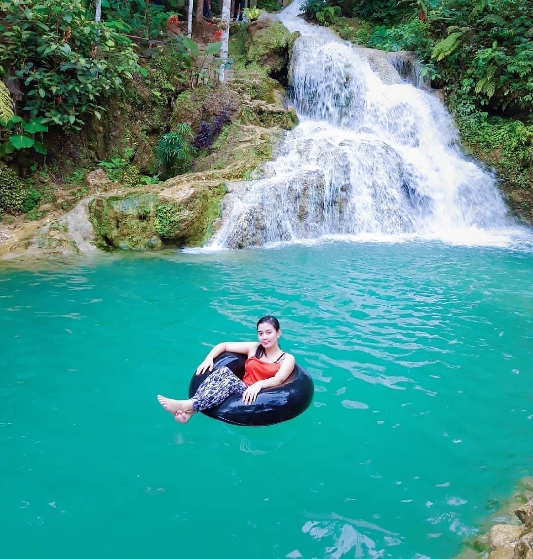 A woman floats on an inflatable ring in the turquoise water with a cascading waterfall in the background at Mudal River Park.