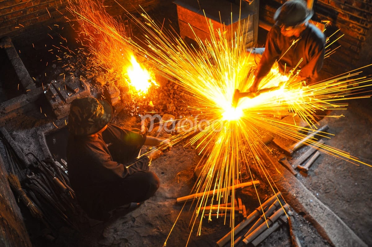 Two craftsmen forging a keris while orange sparks fly from the glowing hot metal in a traditional forge.