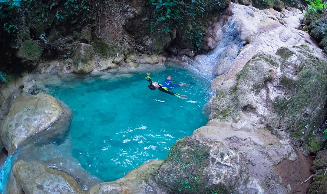 A person swimming in a small, clear, turquoise pool surrounded by rocky terrain and greenery at Mudal River Park.