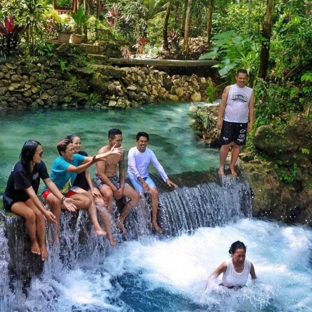 A group of people enjoying the waterfall at Mudal River Park, sitting and standing by the cascading water in a lush green setting.