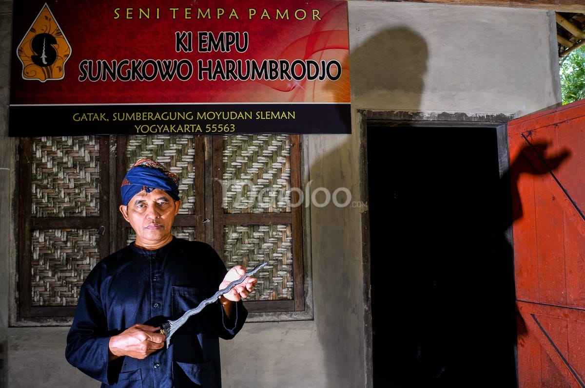 A portrait of Ki Empu Sungkowo Harumbrodjo standing in front of his padepokan, holding a keris.