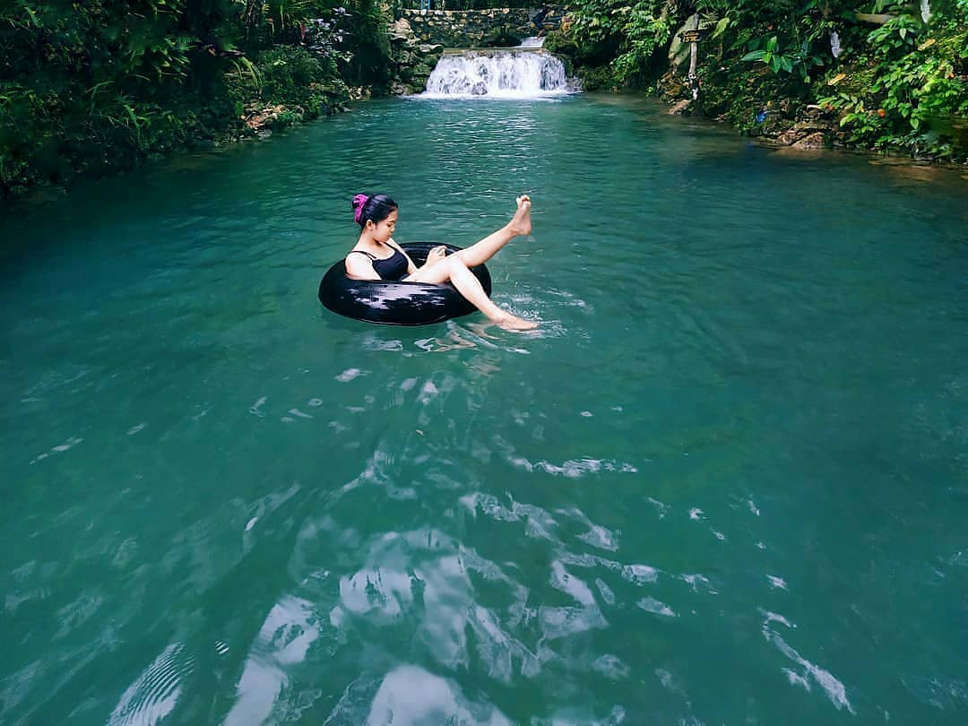 A girl floating on an inflatable ring in the turquoise waters of Mudal River Park with a small waterfall in the background.