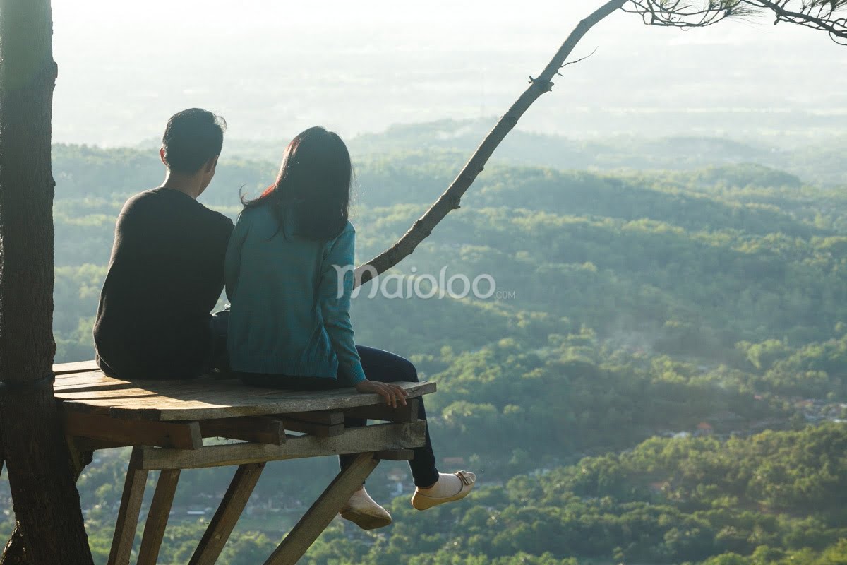 A young couple sits on a wooden viewing platform at Becici Peak, enjoying the expansive view of the green landscape.