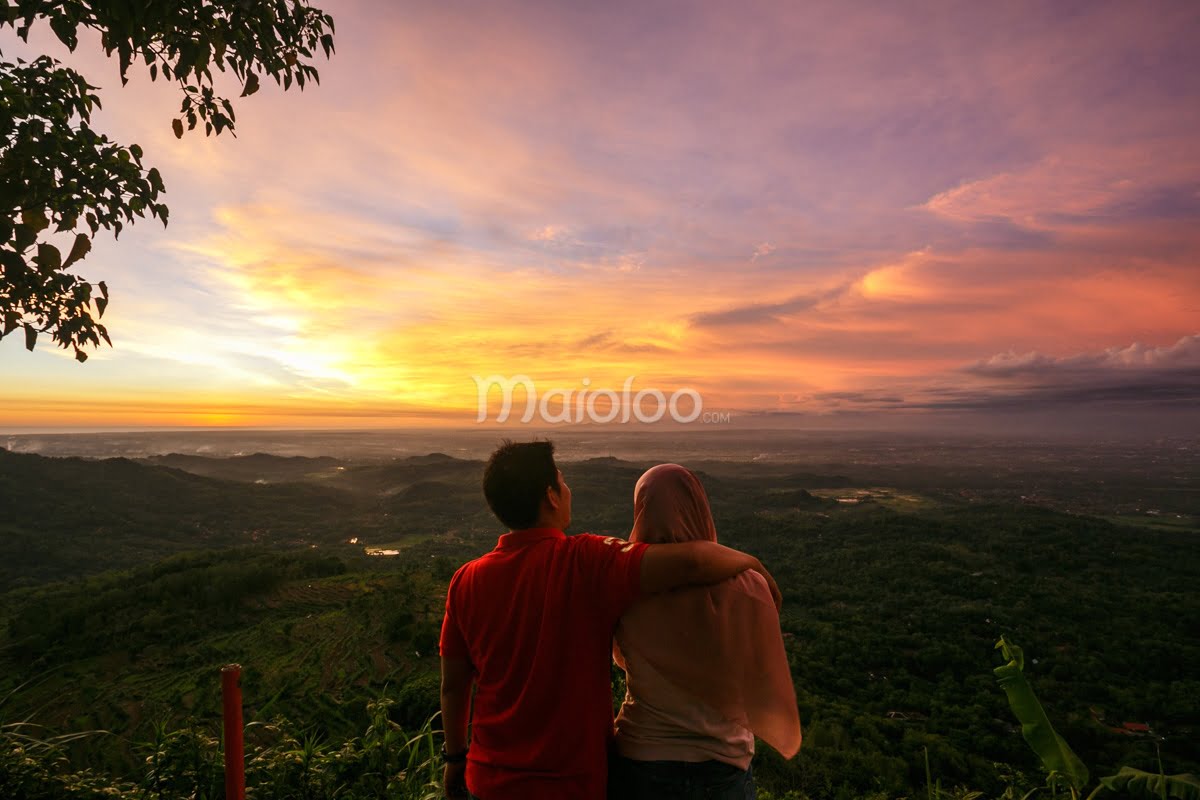 A couple watches a vibrant sunset over a lush green landscape from Becici Peak.