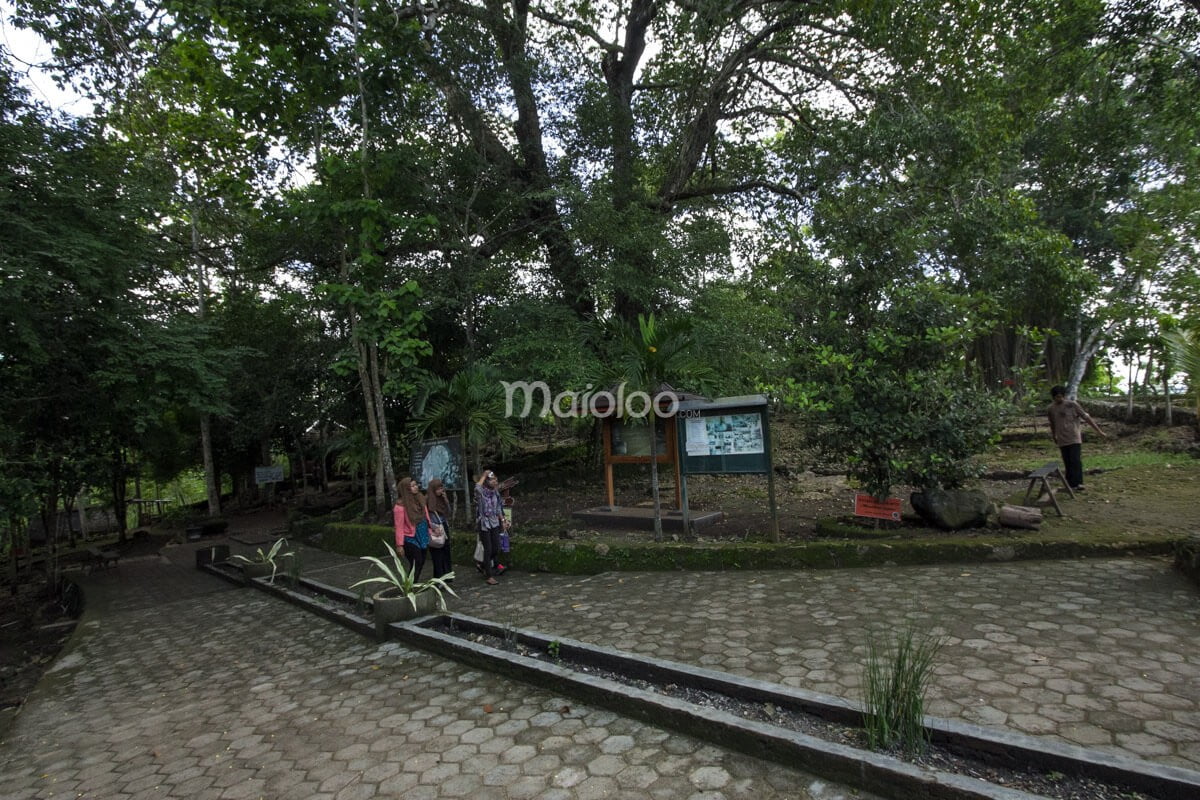 Visitors walking near the entrance area of Rancang Kencono Cave, surrounded by trees and informational boards.