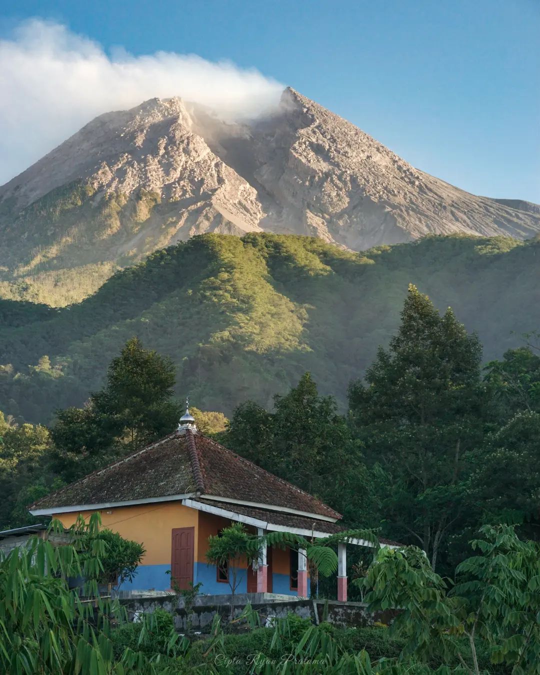 A small mosque surrounded by greenery at Klangon Hill, with Mount Merapi towering in the background under a clear sky.