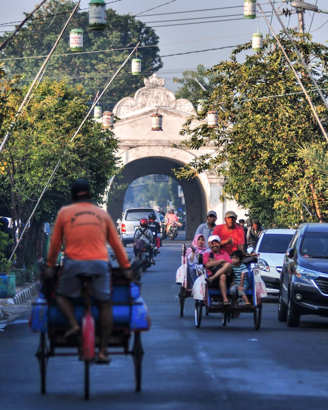 A street view leading to Plengkung Gading in Yogyakarta. The road is busy with motorcycles, cars, and traditional becaks carrying passengers under hanging lanterns. The historical gate is visible in the background.