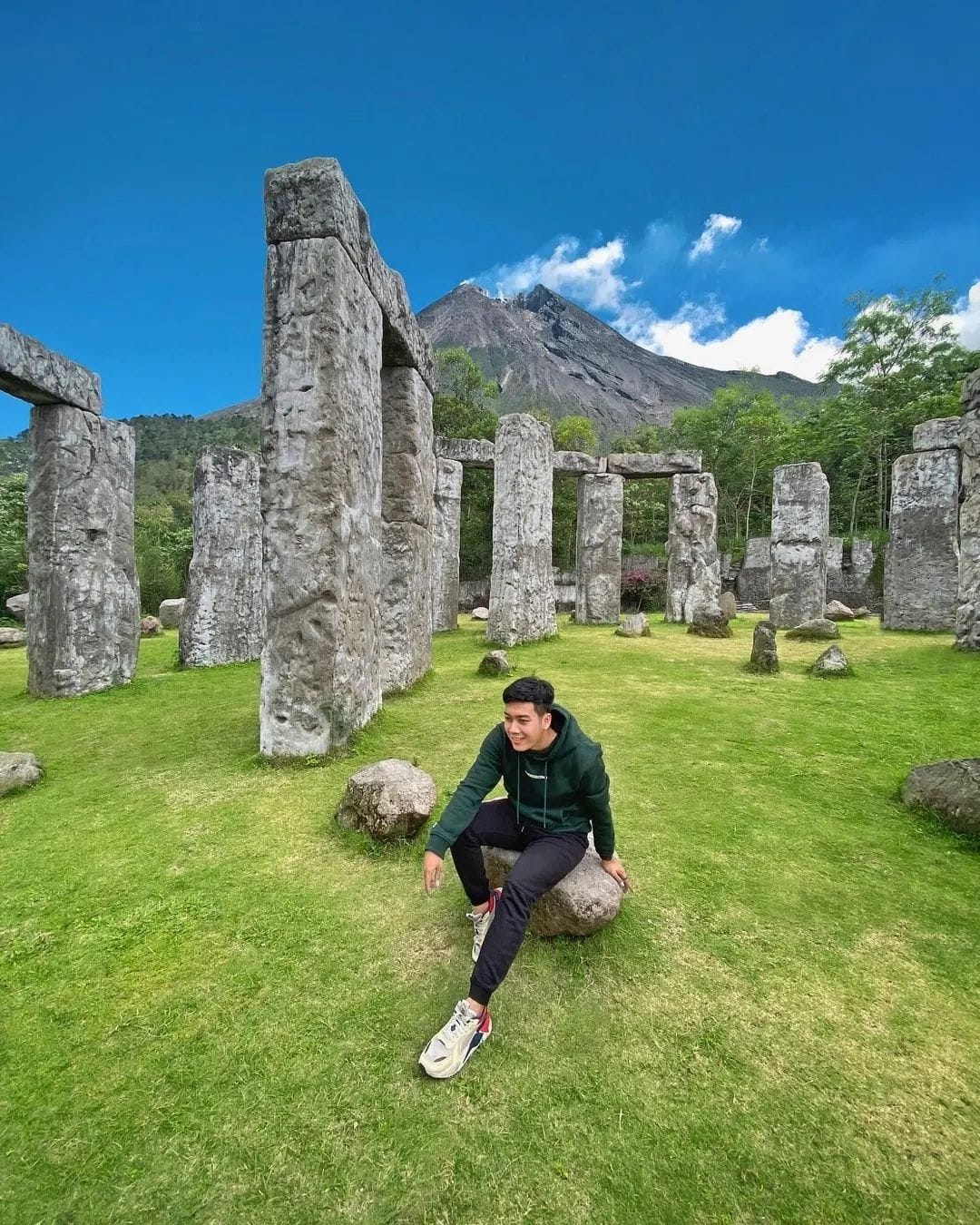 A man sits on a small rock among the stone pillars of Stonehenge Yogyakarta, with the towering peak of Mount Merapi visible in the background under a bright blue sky. The grassy area and stone structures make for an outdoor, scenic setting.