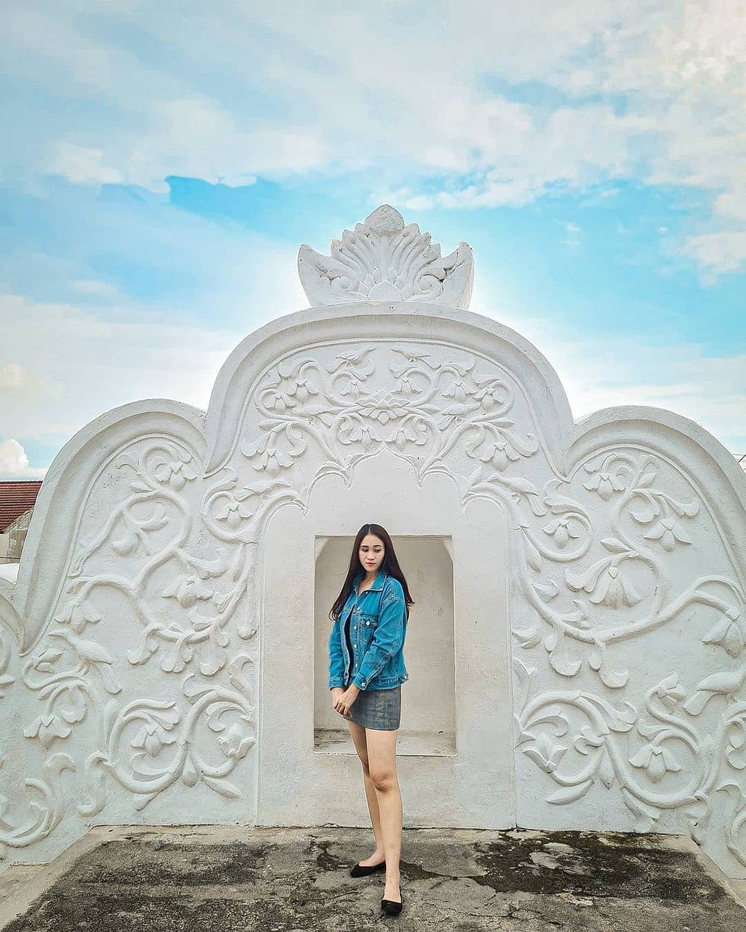 A young woman stands in front of an ornately carved section of Plengkung Gading in Yogyakarta. The white stone carvings feature intricate floral patterns, and the blue sky serves as a backdrop.