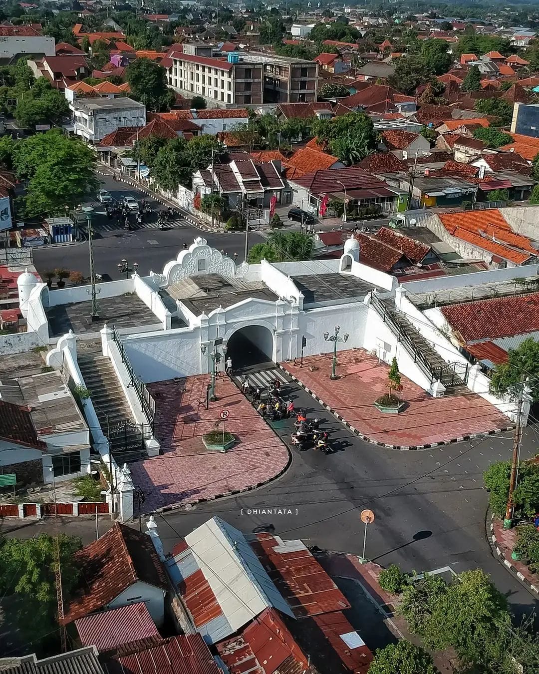 An aerial shot of Plengkung Gading in Yogyakarta. The white southern gate is surrounded by red-roofed houses, modern buildings, and local streets with people riding motorcycles through the gate.