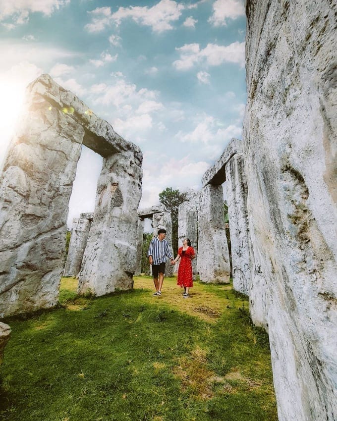 A couple walks hand-in-hand between the large stone pillars of the Stonehenge Yogyakarta replica under a bright, partly cloudy sky. The sun is shining through the stones, casting a soft glow over the scene.