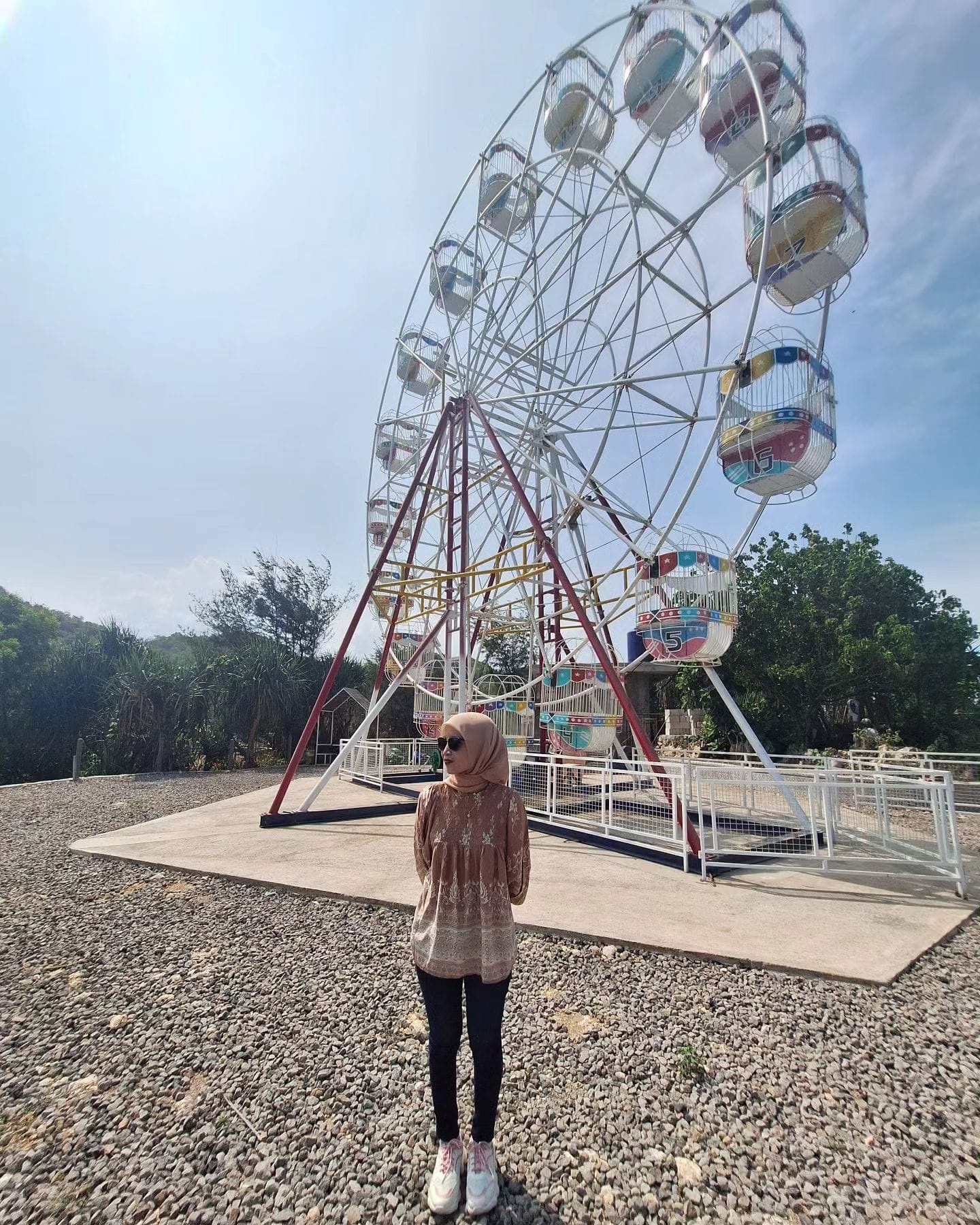 A visitor stands in front of the giant Ferris wheel at Gesing Wonderland. The Ferris wheel features colorful cabins and stands tall under a bright sky, surrounded by greenery and gravel ground.