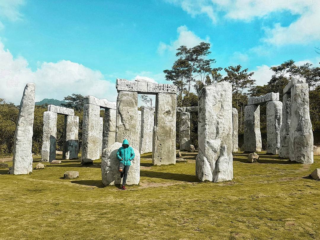 A person in a bright blue hoodie leans against one of the large stone pillars at Stonehenge Yogyakarta. The full-scale replica of Stonehenge stands tall among trees, under a clear blue sky, with paths of green grass surrounding the stone structures.