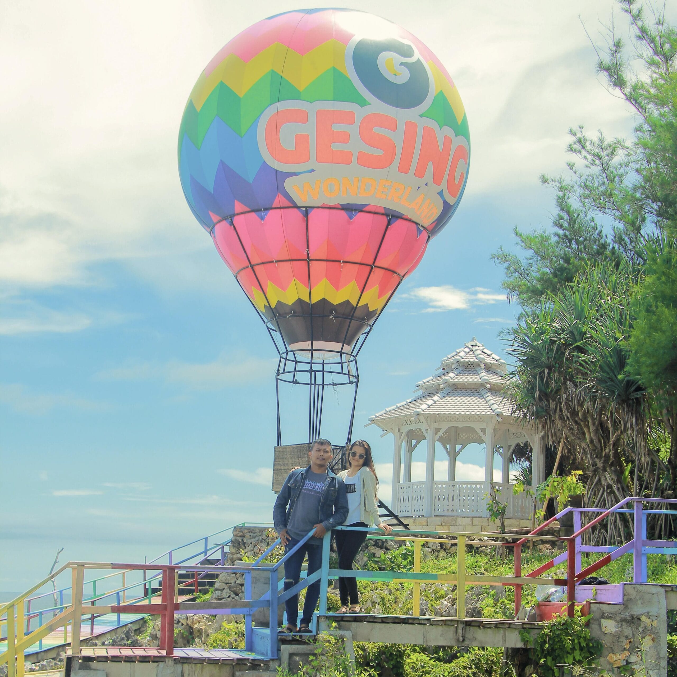 Two visitors stand on a colorful staircase in front of a vibrant hot air balloon display at Gesing Wonderland. A white gazebo is visible in the background, surrounded by greenery and clear blue skies.