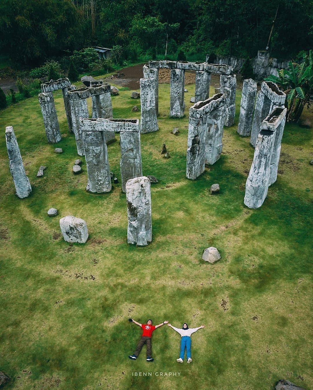 Aerial view of two people lying on the grass with arms outstretched inside the Stonehenge Yogyakarta replica, surrounded by large stone pillars. The green grass and stone formations create a peaceful scene with dense trees in the background.