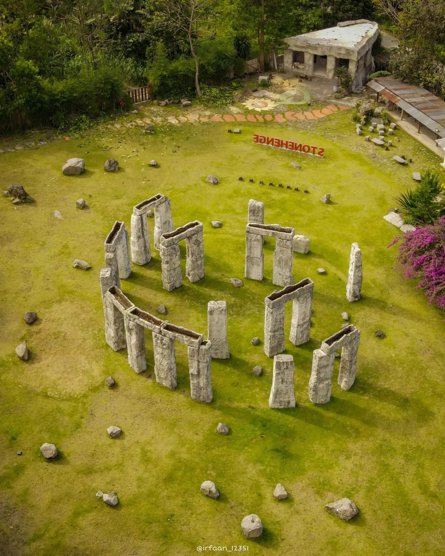 Aerial view of the Stonehenge Yogyakarta replica, showing the stone circle surrounded by green grass and trees. Purple flowers and a stone structure can be seen nearby, with the "Stonehenge" sign visible in the background.