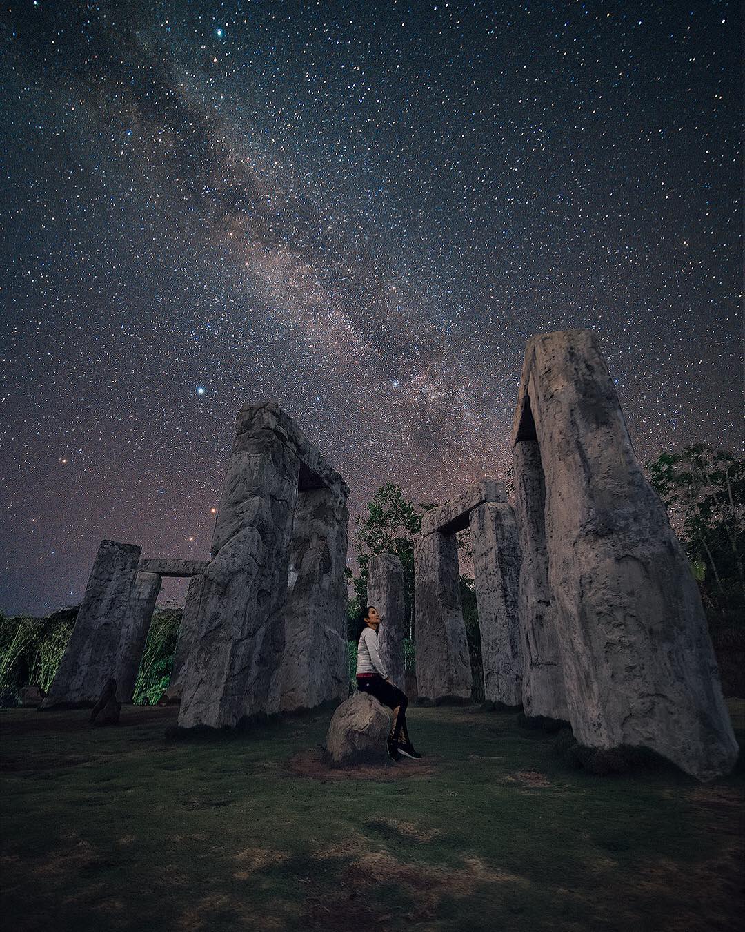 A woman sits on a rock among the towering pillars of the Stonehenge Yogyakarta replica at night, gazing up at the clear sky filled with countless stars and the visible Milky Way.