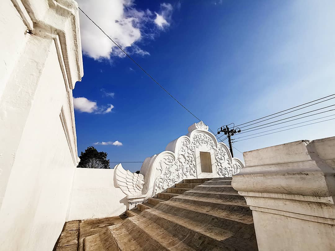 A bright view from the top of Plengkung Gading in Yogyakarta, with a beautifully carved white wall and stone steps leading upward. The sky is clear blue with a few scattered clouds.