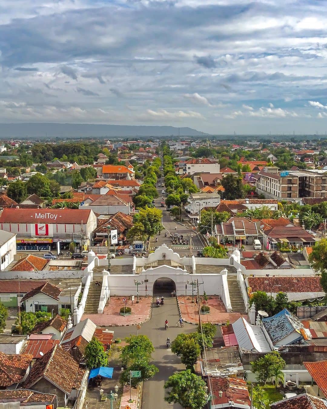 An aerial view of Plengkung Gading, Yogyakarta’s southern gate, showing the gate centered among red-roofed buildings. A long street lined with trees stretches out into the city, with cloudy skies in the background.