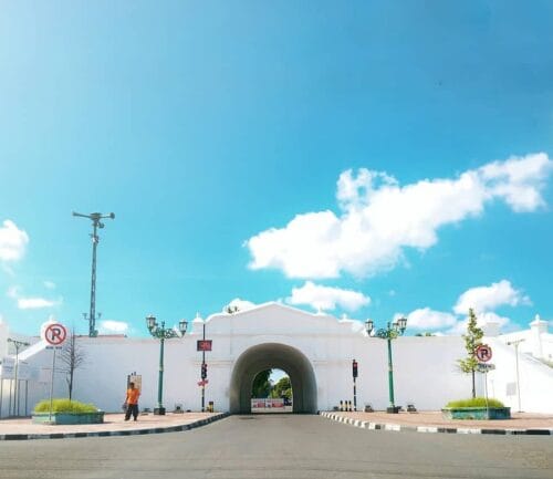 A bright day view of Plengkung Gading, the southern gate of Yogyakarta Palace. The white gate stands under a blue sky with a few clouds. A pedestrian can be seen walking near the sidewalk, and a siren tower stands beside the gate.