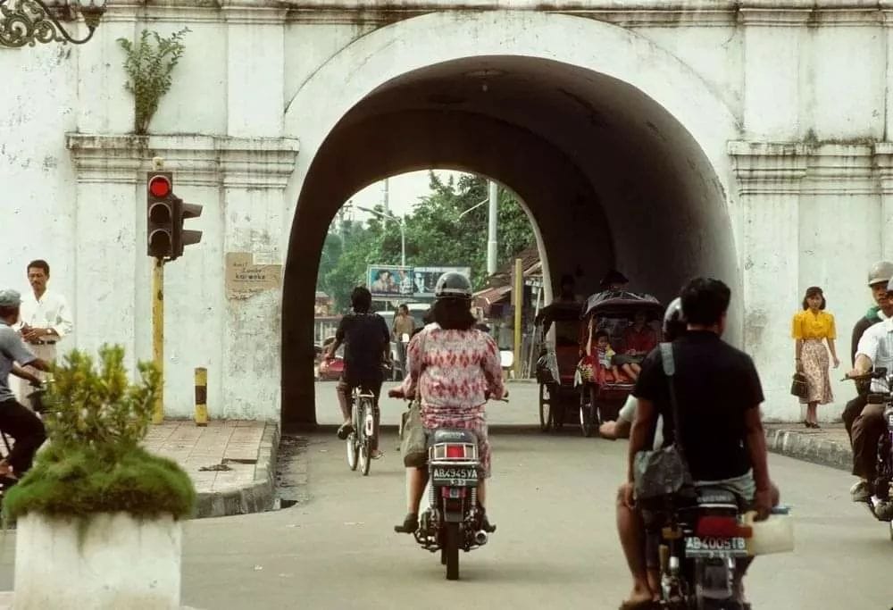 A nostalgic street scene from Yogyakarta in 1992 showing locals passing through Plengkung Gading on bicycles, motorcycles, and a traditional becak (pedicab). A traffic light and pedestrians are visible near the gate.