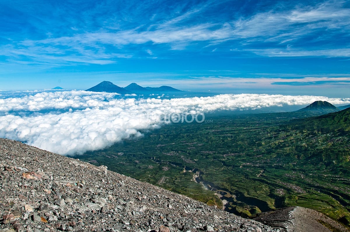 A panoramic view from Pasar Bubrah on Mount Merapi, showing distant mountains rising above a blanket of clouds and the green landscape below.