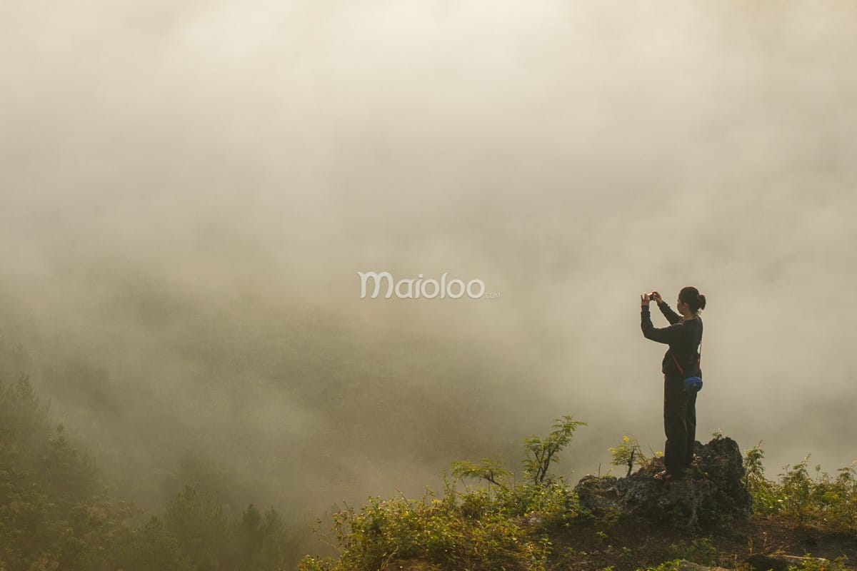 A person stands on a rock at Panguk Kediwung Hill, capturing a photo of the clouds with a smartphone.