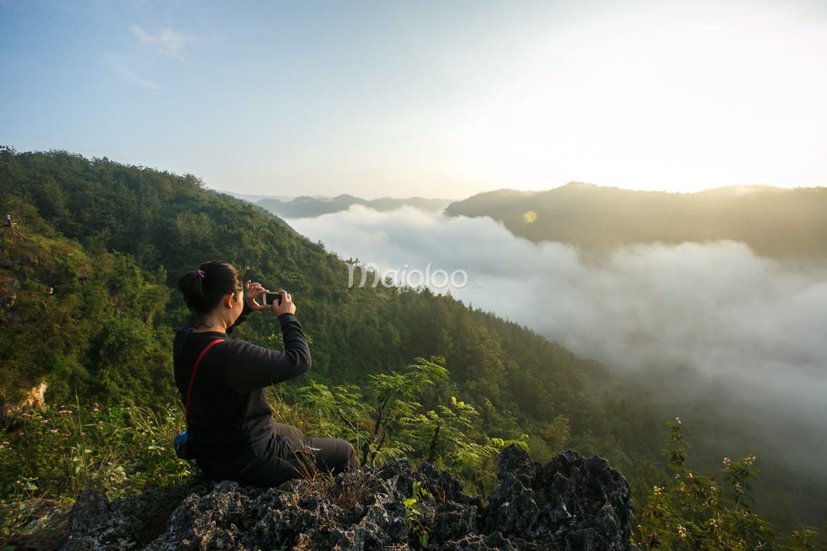 A person sits on a rocky ledge taking photos of the sunrise and clouds at Panguk Kediwung Hill.