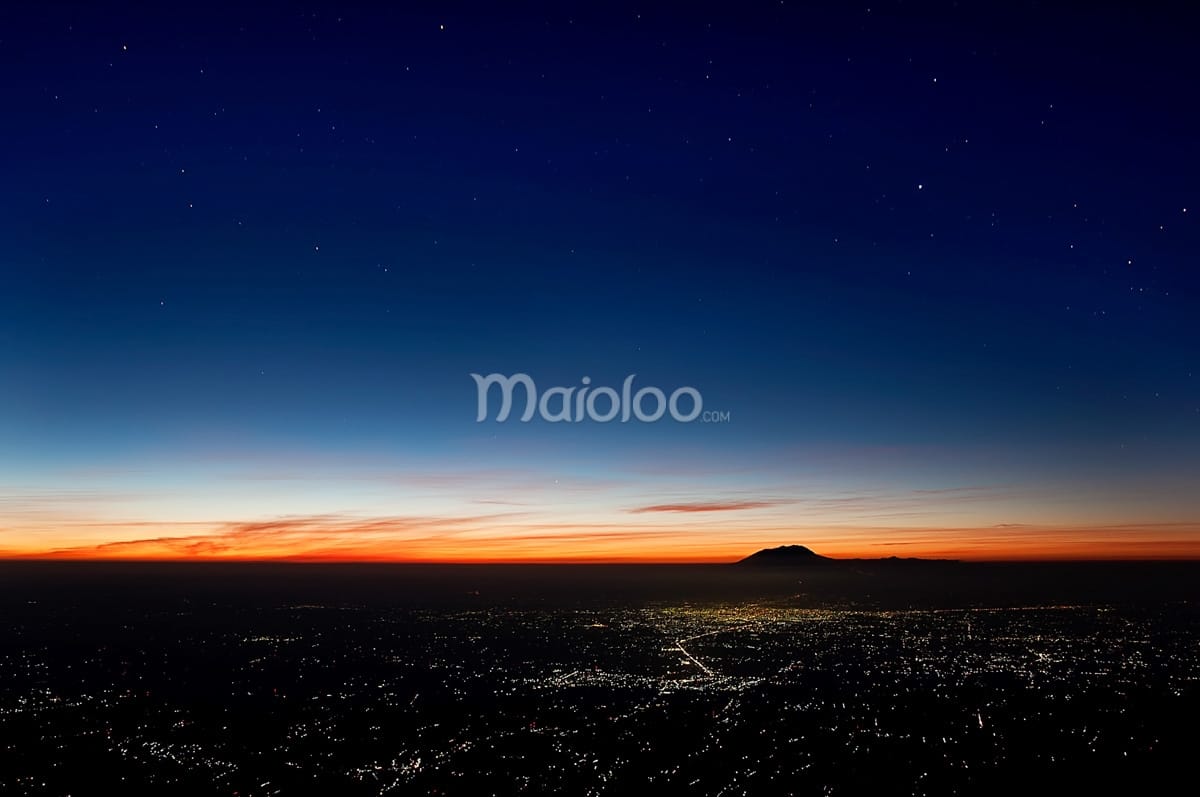 A view of the city lights of Surakarta at night, seen from the hiking trail on Mount Merapi with a colorful horizon and starry sky.