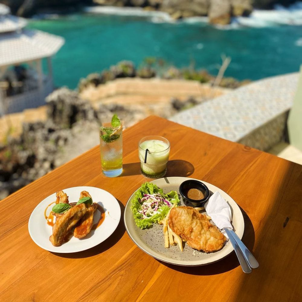 A meal featuring fried fish, salad, French fries, and chicken wings is laid on a wooden table with refreshing drinks. The food is served with a beautiful ocean view in the background at Gesing Wonderland.