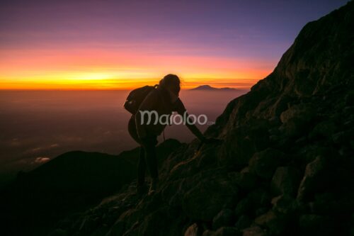 A hiker climbs a rocky path on Mount Merapi during sunrise, with a colorful sky in the background.