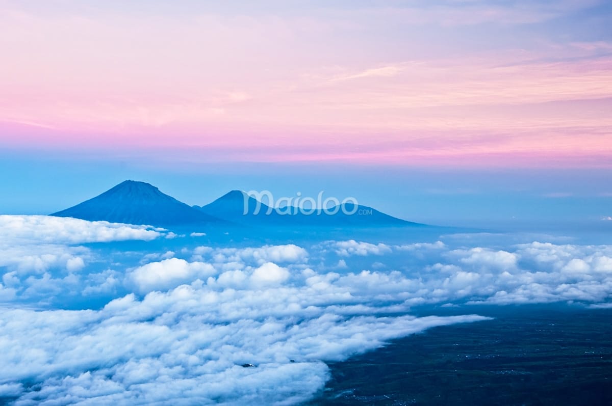 A view of Mount Sumbing, Mount Sindoro, and the Dieng Plateau rising above a blanket of clouds with a colorful dawn sky.