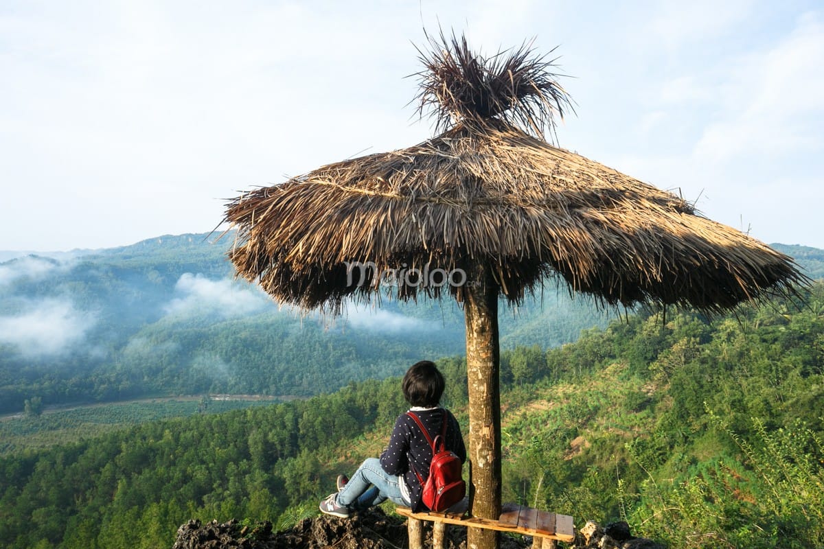 A person sits on a bench under a straw hut, overlooking green hills and clouds at Panguk Kediwung Hill.