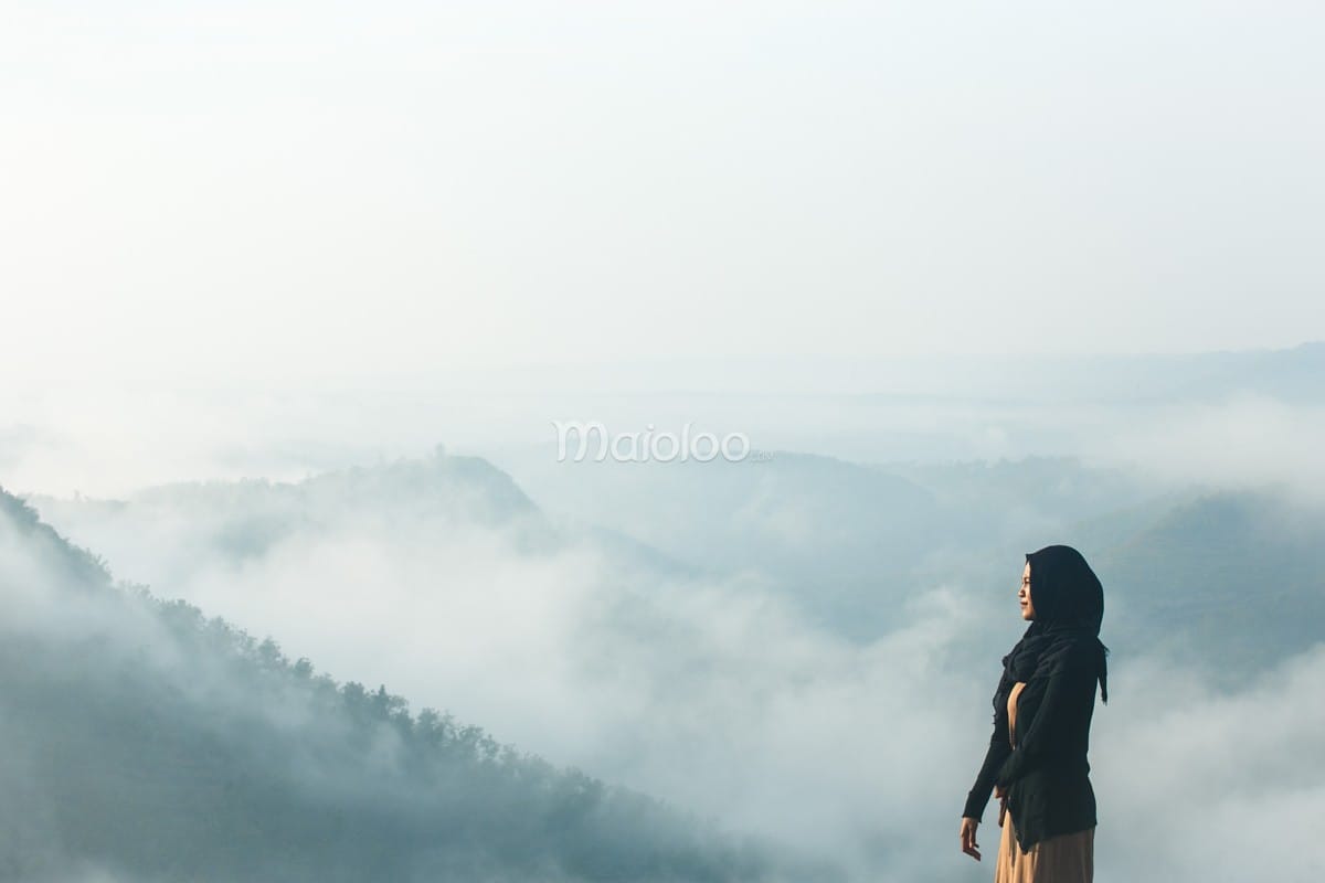 A person looks out over a sea of clouds at Panguk Kediwung Hill, with mist-covered hills stretching into the distance.