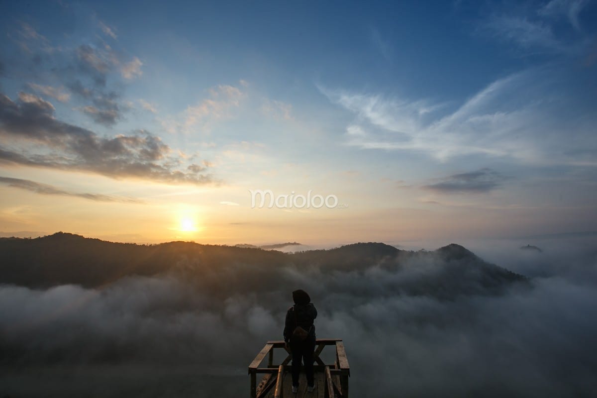 A person stands on a viewing platform at Panguk Kediwung Hill, admiring the sunrise above a sea of clouds.