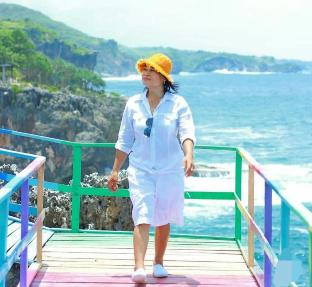 A visitor wearing a yellow hat and white outfit strolls along the colorful Rainbow Bridge at Gesing Wonderland. The bridge overlooks a beautiful ocean with rocky cliffs in the background.