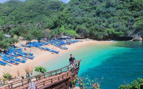 A view of Gesing Beach with turquoise waters and a sandy shore lined with blue fishing boats. A person is standing on a wooden deck overlooking the beach, surrounded by green hills.