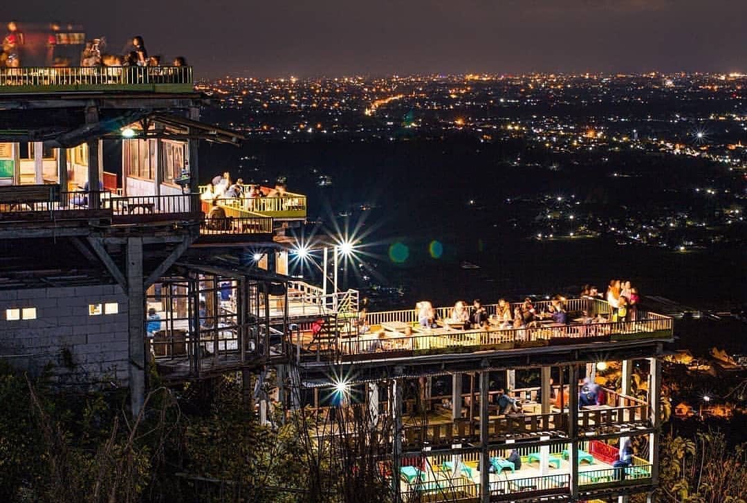 People enjoying the night view from a brightly lit café terrace at Bukit Bintang, with Yogyakarta's twinkling city lights visible in the background.