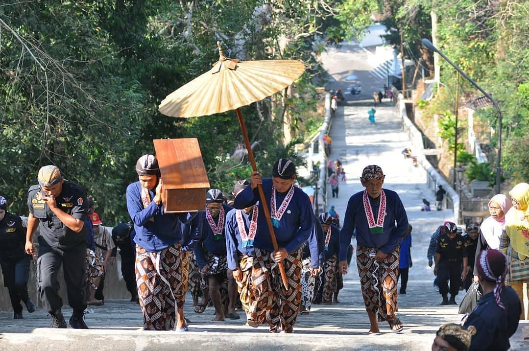 A group of people in traditional Javanese clothing participate in the Nguras Enceh ritual, carrying sacred items while climbing the stone steps of Imogiri Royal Cemetery.