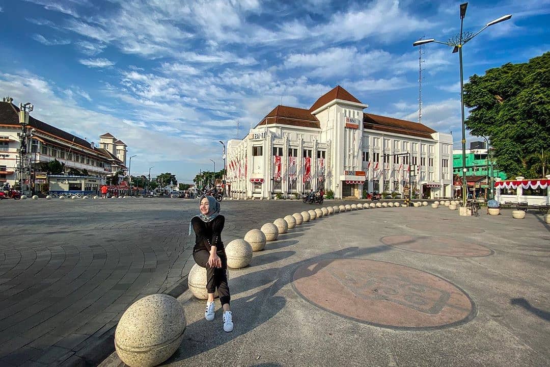 A woman sitting on a stone bollard in front of the iconic white BNI building at Yogyakarta Kilometer Zero. The sky is blue, and the historic building and surrounding area are clearly visible in the background.