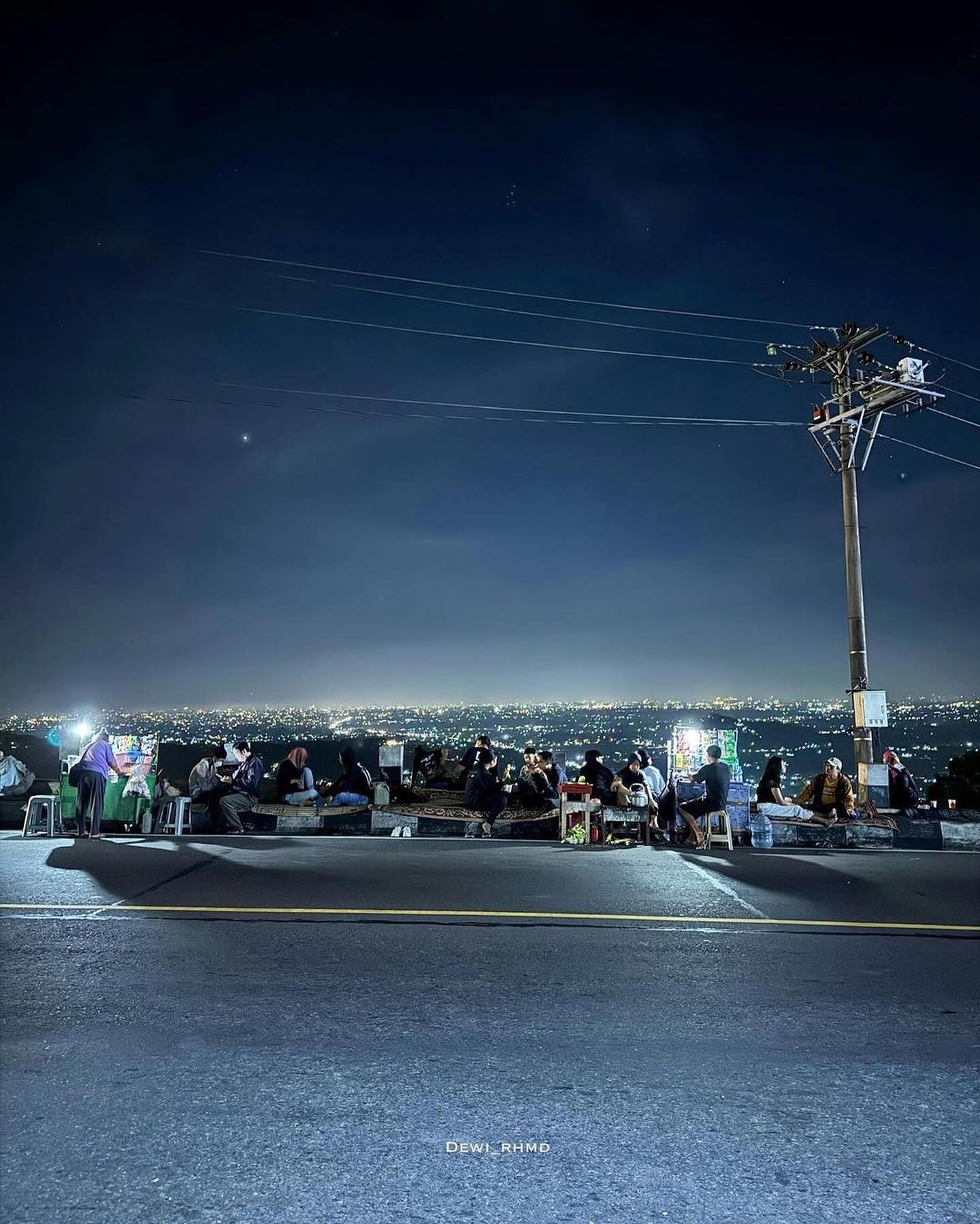 Street vendors selling roasted corn and instant drinks to visitors sitting along the roadside at Bukit Bintang, with Yogyakarta's city lights glowing in the distance at night.