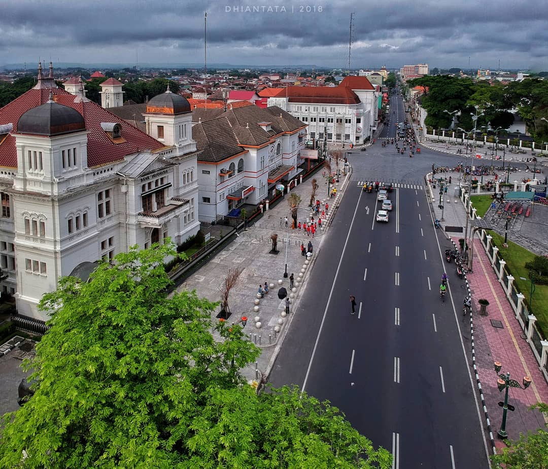 An aerial view of Yogyakarta Kilometer Zero, featuring the historic BNI building, Yogyakarta Post Office, and surrounding streets. The roads are lined with trees and buildings, with a mix of traffic and pedestrians.