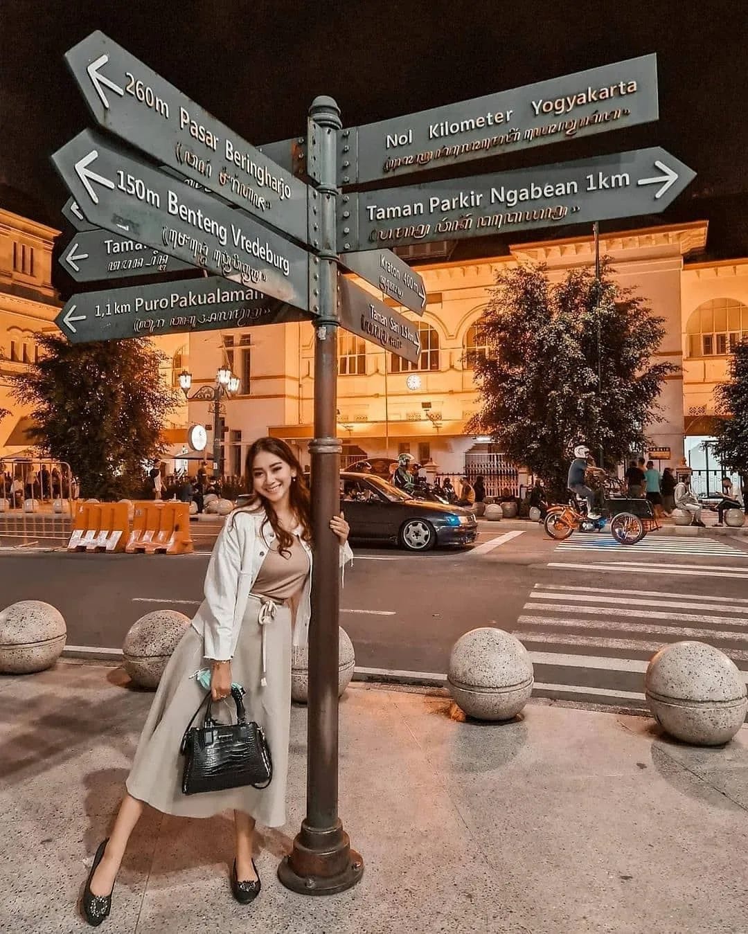 A young woman standing next to a signpost at Yogyakarta Kilometer Zero at night. The signs point to various nearby attractions while traffic and pedestrians are visible in the background.