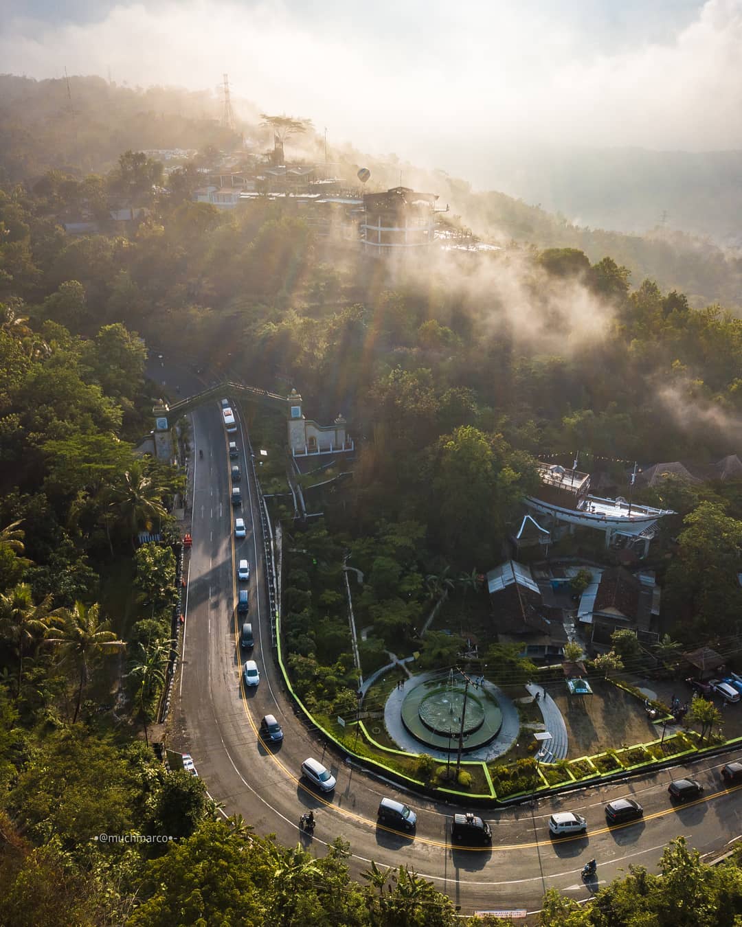 An aerial view of a winding road surrounded by lush green trees and mist near Bukit Bintang, with cars driving around a curve and buildings nestled along the hillside.