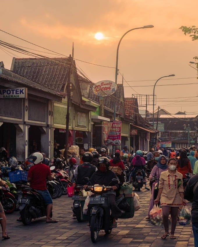 Busy street scene at Pasar Kotagede during sunset, with people walking, riding motorcycles, and shopping in a lively atmosphere.