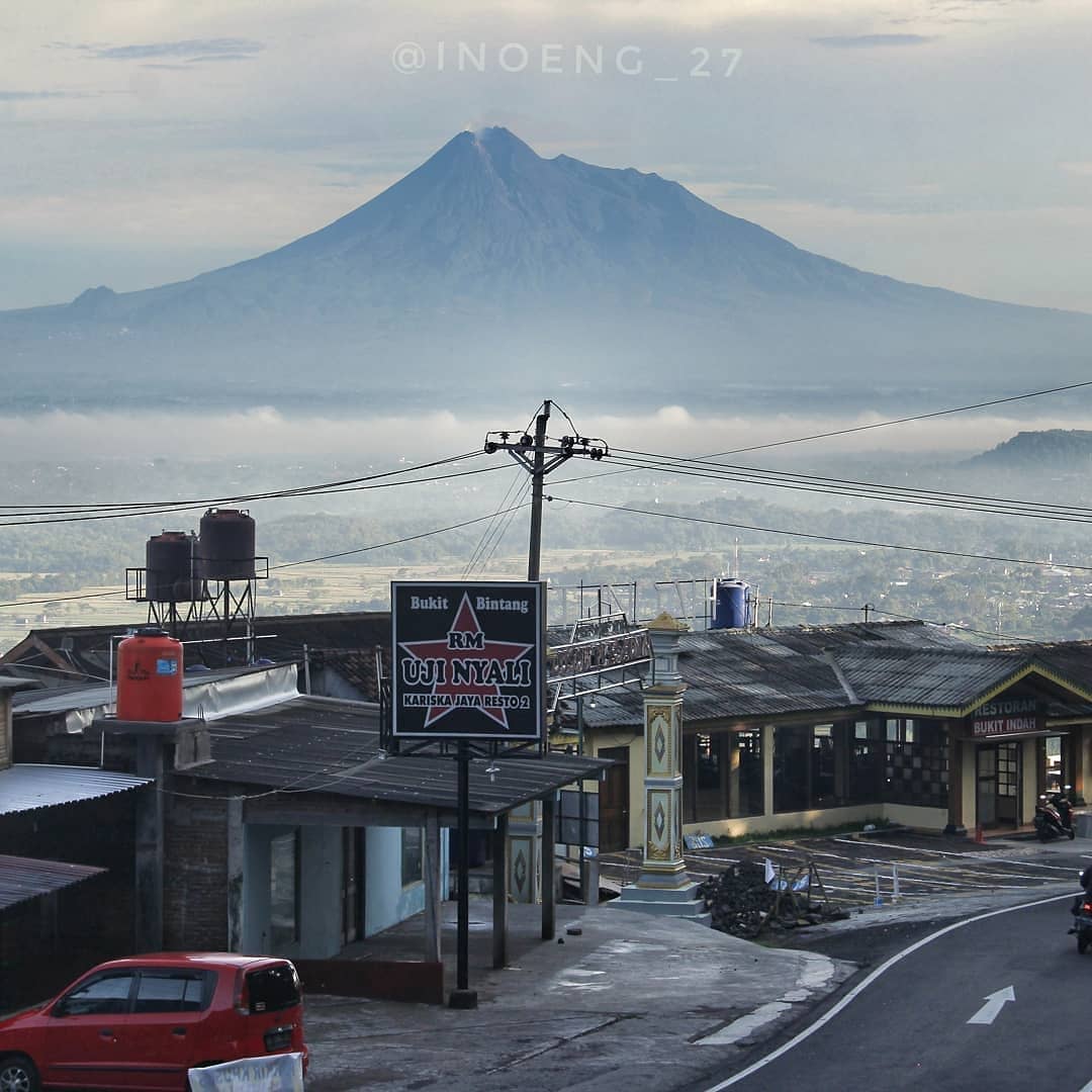 Mount Merapi rises in the background, seen from a quiet road in Bukit Bintang with local restaurants and a hillside view.