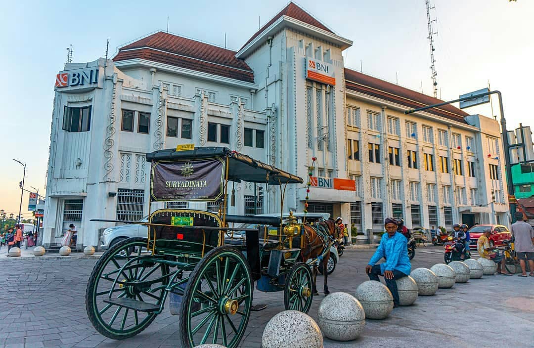 A traditional horse-drawn carriage, or "andong," parked in front of the historic BNI building at Yogyakarta Kilometer Zero. The carriage driver is sitting nearby, while people can be seen walking and riding motorbikes in the background.